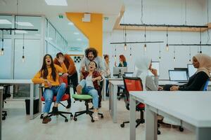 Team building and office fun. Young cheerful businesspeople in smart casual wear having fun while racing on office chairs and smiling. photo