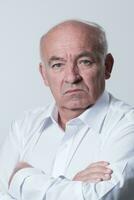 Confident senior man in white shirt crossing hands on chest and looking at camera while standing against gray background. Self confident senior isolated white studio shoot. photo