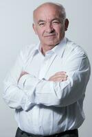 Confident senior man in white shirt crossing hands on chest and looking at camera while standing against gray background. Self confident senior isolated white studio shoot. photo