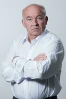 Confident senior man in white shirt crossing hands on chest and looking at camera while standing against gray background. Self confident senior isolated white studio shoot. photo