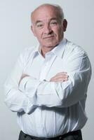 Confident senior man in white shirt crossing hands on chest and looking at camera while standing against gray background. Self confident senior isolated white studio shoot. photo