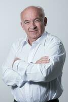 Confident senior man in white shirt crossing hands on chest and looking at camera while standing against gray background. Self confident senior isolated white studio shoot. photo