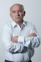 Confident senior man in white shirt crossing hands on chest and looking at camera while standing against gray background. Self confident senior isolated white studio shoot. photo