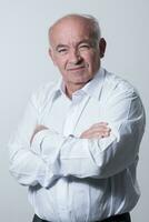 Confident senior man in white shirt crossing hands on chest and looking at camera while standing against gray background. Self confident senior isolated white studio shoot. photo