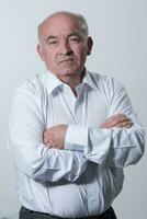 Confident senior man in white shirt crossing hands on chest and looking at camera while standing against gray background. Self confident senior isolated white studio shoot. photo