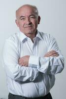 Confident senior man in white shirt crossing hands on chest and looking at camera while standing against gray background. Self confident senior isolated white studio shoot. photo