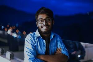 un indio hombre con lentes y un azul camisa mira alrededor el ciudad a noche. en el antecedentes de el noche calle de el ciudad foto