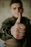 The soldier makes a gesture of success with his hand. A soldier in full war gear stands in front of a stone wall and shows the ok sign with his finger photo