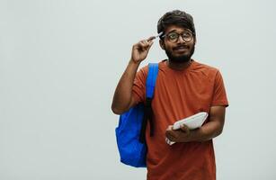 confuso indio estudiante con azul mochila, lentes y cuaderno posando en gris antecedentes. el concepto de educación y enseñanza. hora a Vamos espalda a colegio foto