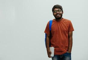 indio estudiante con azul mochila, lentes y cuaderno posando en gris antecedentes. el concepto de educación y enseñanza. hora a Vamos espalda a colegio foto