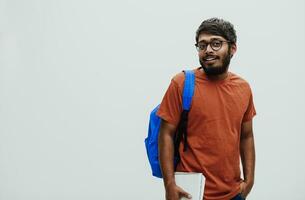 indio estudiante con azul mochila, lentes y cuaderno posando en gris antecedentes. el concepto de educación y enseñanza. hora a Vamos espalda a colegio foto