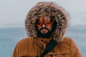 disparo a la cabeza foto de un hombre en un frío Nevado zona vistiendo un grueso marrón invierno chaqueta, nieve gafas de protección y guantes. vida en frío regiones de el país.