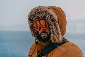 disparo a la cabeza foto de un hombre en un frío Nevado zona vistiendo un grueso marrón invierno chaqueta, nieve gafas de protección y guantes. vida en frío regiones de el país.