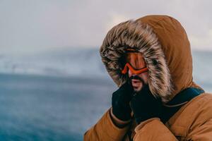 disparo a la cabeza foto de un hombre en un frío Nevado zona vistiendo un grueso marrón invierno chaqueta, nieve gafas de protección y guantes. vida en frío regiones de el país.