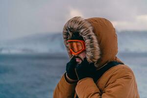 disparo a la cabeza foto de un hombre en un frío Nevado zona vistiendo un grueso marrón invierno chaqueta, nieve gafas de protección y guantes. vida en frío regiones de el país.