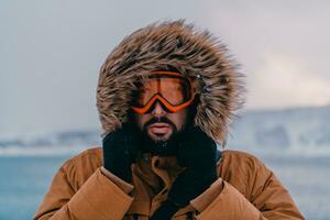 disparo a la cabeza foto de un hombre en un frío Nevado zona vistiendo un grueso marrón invierno chaqueta, nieve gafas de protección y guantes. vida en frío regiones de el país.