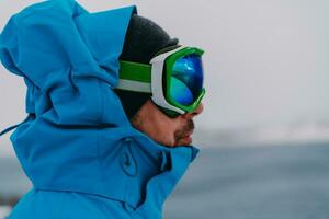 disparo a la cabeza foto de un hombre en un frío Nevado zona vistiendo un grueso azul invierno chaqueta, nieve gafas de protección y guantes. vida en frío regiones de el país.