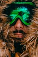 Headshot photo of a man in a cold snowy area wearing a thick brown winter jacket, snow goggles and gloves. Life in cold regions of the country.