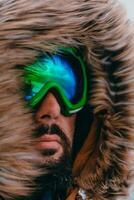 Headshot photo of a man in a cold snowy area wearing a thick brown winter jacket, snow goggles and gloves. Life in cold regions of the country.