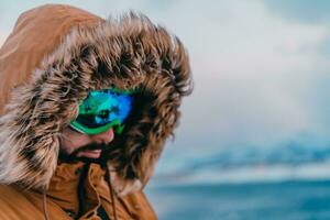 disparo a la cabeza foto de un hombre en un frío Nevado zona vistiendo un grueso marrón invierno chaqueta, nieve gafas de protección y guantes. vida en frío regiones de el país.