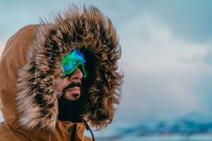 disparo a la cabeza foto de un hombre en un frío Nevado zona vistiendo un grueso marrón invierno chaqueta, nieve gafas de protección y guantes. vida en frío regiones de el país.