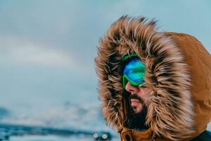 disparo a la cabeza foto de un hombre en un frío Nevado zona vistiendo un grueso marrón invierno chaqueta, nieve gafas de protección y guantes. vida en frío regiones de el país.