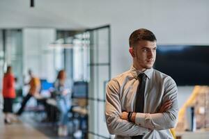 A young business leader stands with crossed arms in a modern office hallway, radiating confidence and a sense of purpose, embodying a dynamic and inspirational presence. photo