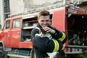 Close-up portrait of heroic fireman in protective suit and red helmet holds saved cat in his arms. Firefighter in fire fighting operation. photo