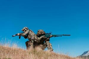 A sniper team squad of soldiers is going undercover. Sniper assistant and team leader walking and aiming in nature with yellow grass and blue sky. Tactical camouflage uniform. photo