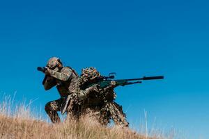 A sniper team squad of soldiers is going undercover. Sniper assistant and team leader walking and aiming in nature with yellow grass and blue sky. Tactical camouflage uniform. photo