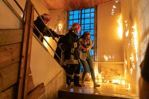 Brave Fireman Descends Stairs of a Burning Building and Holds Saved Girl in His Arms. Open fire and one Firefighter in the Background. photo