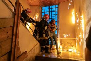 Brave Fireman Descends Stairs of a Burning Building and Holds Saved Girl in His Arms. Open fire and one Firefighter in the Background. photo