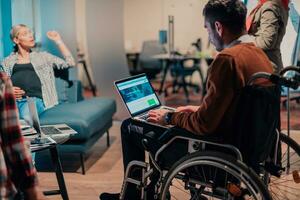 un empresario en un silla de ruedas en un moderno trabajo colaborativo oficina espacio trabajando en ordenador portátil. foto