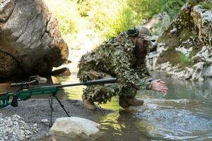 soldado en un camuflaje traje uniforme Bebiendo Fresco agua desde el río. militar francotirador rifle en el lado. foto