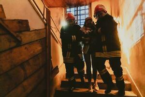 Brave Fireman Descends Stairs of a Burning Building and Holds Saved Girl in His Arms. Open fire and one Firefighter in the Background. photo