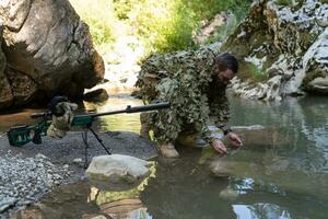 soldado en un camuflaje traje uniforme Bebiendo Fresco agua desde el río. militar francotirador rifle en el lado. foto