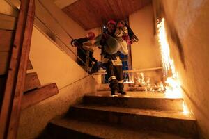 valiente bombero desciende escalera de un ardiente edificio y sostiene salvado niña en su brazos. abierto fuego y uno bombero en el antecedentes. foto