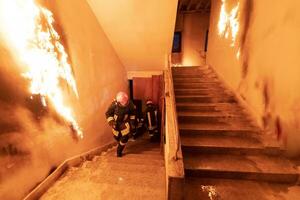 Brave Fireman Descends Stairs of a Burning Building and Holds Saved Girl in His Arms. Open fire and one Firefighter in the Background. photo