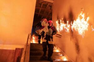 Brave Fireman Descends Stairs of a Burning Building and Holds Saved Girl in His Arms. Open fire and one Firefighter in the Background. photo