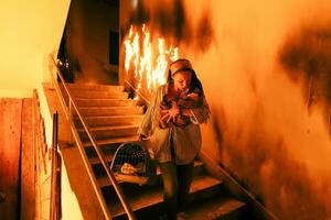 Brave Fireman Descends Stairs of a Burning Building and Holds Saved Girl in His Arms. Open fire and one Firefighter in the Background. photo