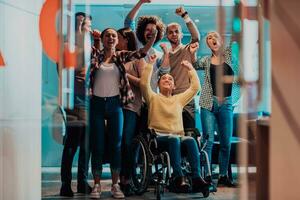 Photo of business women in wheelchairs with their hands raised in the air with their colleagues, together celebrating business success