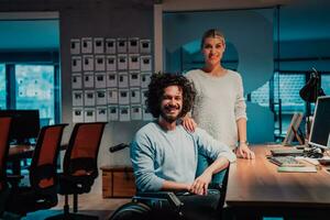 A businessman in a wheelchair in a modern coworking office space working with a female colleague late at nigh. Colleagues in the background photo