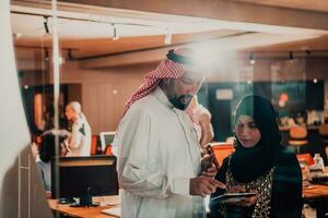 retrato foto de árabe líder empresario con musulmán hijab mujer que se discute negocio proyectos mientras utilizando tabletas y teléfono inteligente en moderno vaso oficina