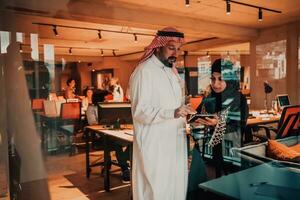 Portrait photo of Arab leader businessman with muslim hijab woman discussing business projects while using tablets and smartphone in modern glass office