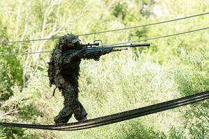 un militar hombre o airsoft jugador en un camuflaje traje furtivo el cuerda puente y objetivos desde un francotirador rifle a el lado o a objetivo foto