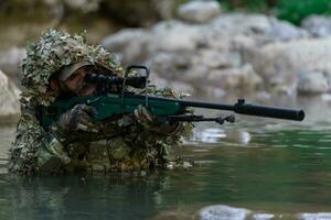 A military man or airsoft player in a camouflage suit sneaking the river and aims from a sniper rifle to the side or to target. photo