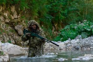 A military man or airsoft player in a camouflage suit sneaking the river and aims from a sniper rifle to the side or to target. photo