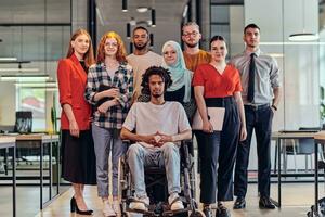 A diverse group of young business people congregates within a modern startup's glass-enclosed office, featuring inclusivity with a person in a wheelchair, an African American young man , and a hijab-clad woman, showcasing a dynamic blend of innovation and photo