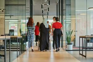 A diverse group of business people walking a corridor in the glass-enclosed office of a modern startup, including a person in a wheelchair and a woman wearing a hijab, showing a dynamic mix of innovation and unity. photo