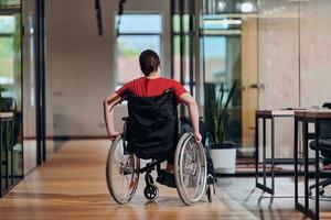 A modern young businesswoman in a wheelchair is surrounded by an inclusive workspace with glass-walled offices, embodying determination and innovation in the business world photo